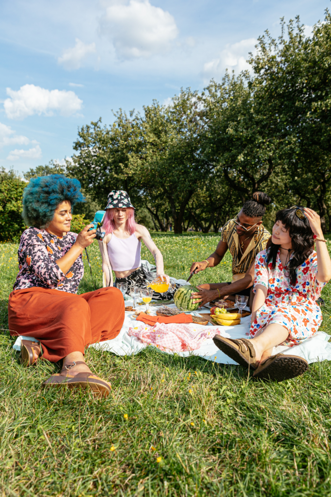 friends enjoying a picnic in a local park. Diversity is represented with a black woman with blue hair, an Asian woman, a black man, and a white woman with pink hair. 