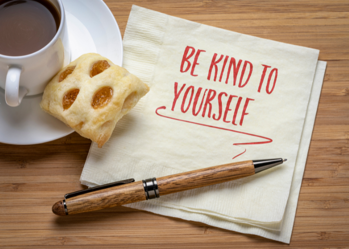 Be kind to yourself is written on a napkin. There is also a cup of coffee and a pen. The reminder is the be kind to yourself when you decide where to live. 