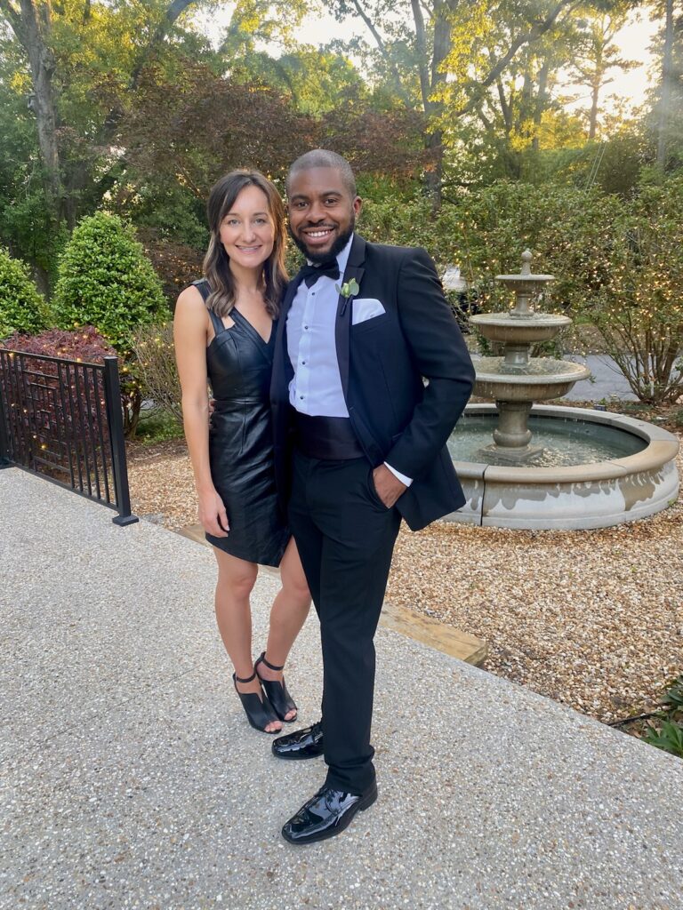 founder Brit Suits of Choosewheretolive.com stands next to her boyfriend. They are wearing formal attire and happily standing next to each other after meeting on a dating app. Brit is happy that her life goals and how to decide where to live are combined. 