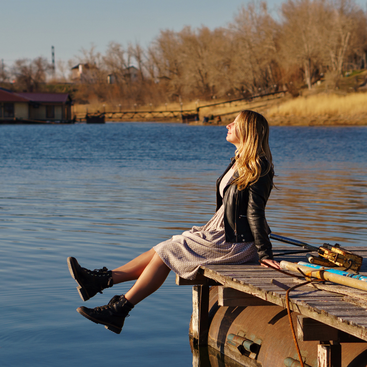 White girl sitting on lake with leather jacket, a dress, and boots on. She has her face titled towards the sun happy that she has chosen to love where she lives! 